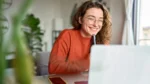 Woman sits at laptop smiling at screen while holding a pen