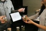 A dispensary employee assists a customer at the counter, holding a labeled cannabis container while verifying information on a digital point-of-sale screen.