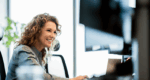Administrator smiling at her computer while reviewing assessments in a modern office environment.