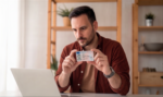 A person sitting at a laptop holding up a government-issued ID card to the camera during an online exam identity verification check
