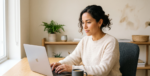 A candid, medium shot of a focused Hispanic woman in her 30s with curly dark hair, wearing a cream sweater, seated at a light wood desk and typing on a silver laptop. The desk is next to a large window providing soft natural light. In the background, there's a simple shelf with plants. A grey coffee mug is on the table.