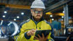 Manufacturing worker in hard hat and high-visibility jacket completing online safety training on a tablet inside an industrial facility