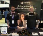 Integrity Advocate CEO Brandon Smith (right) and Blake Donaldson (left) standing at their exhibition booth during the 2026 International e-Assessment Conference in London. They are wearing matching black branded shirts in front of a backdrop that reads 